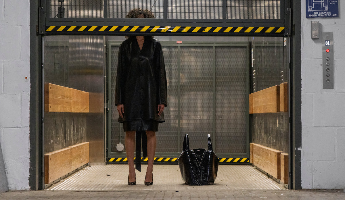 Woman standing in an industrial elevator with the LA MAMA LM1 luxury handbag in Black Lacquer Croco on the floor.