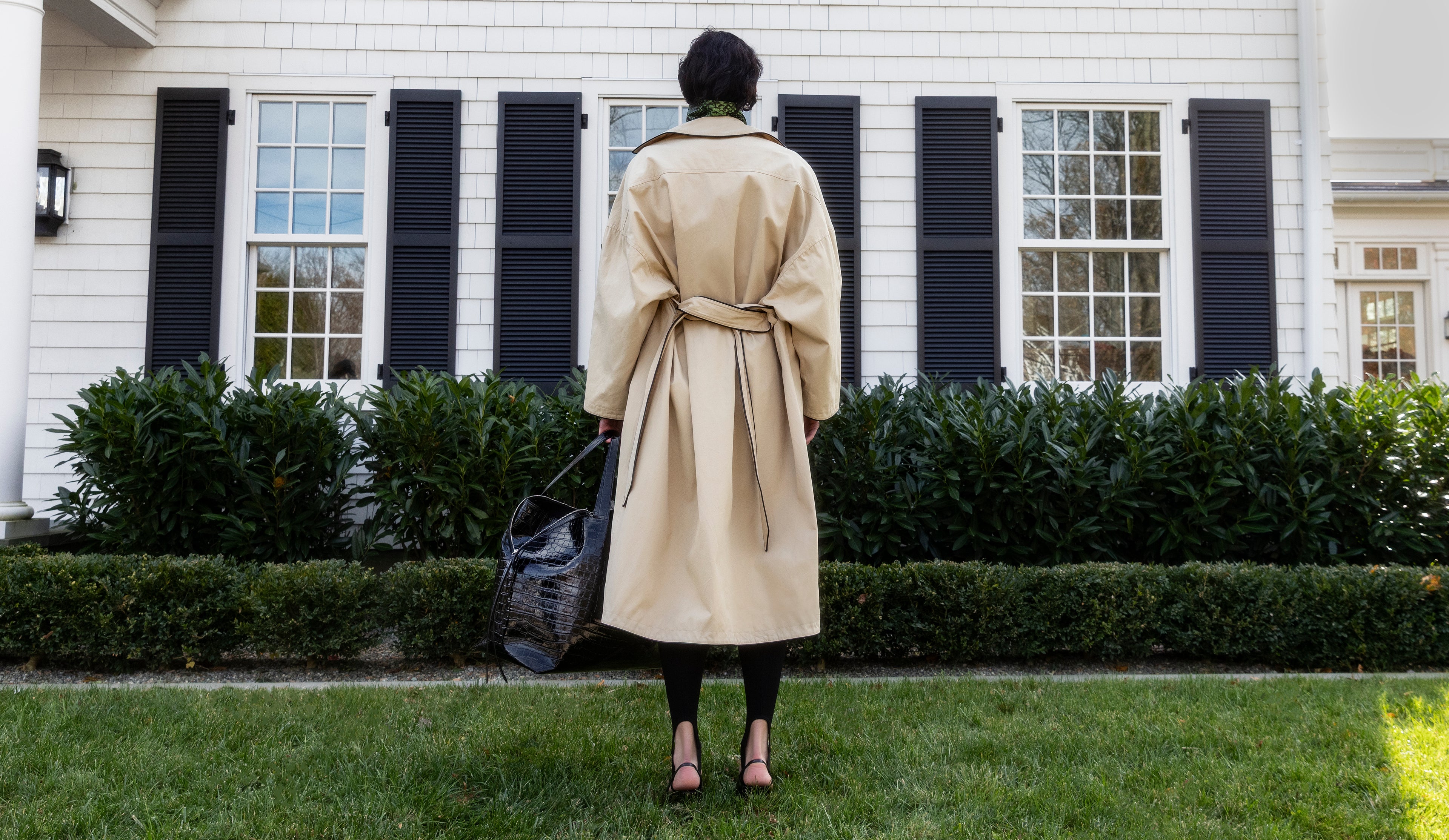 Woman standing in front of a colonial style house carrying the LA MAMA LM1 luxury handbag in Black Lacquer Croco in her hand.