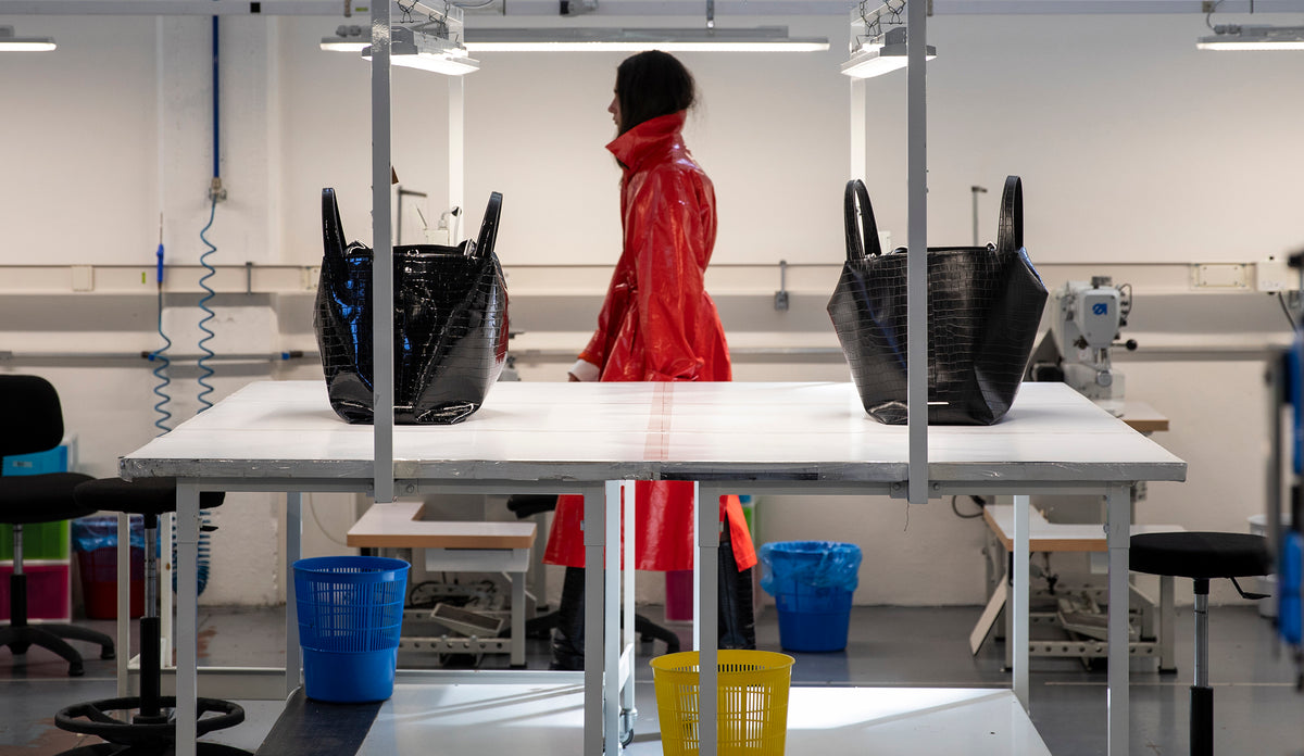 Woman walking in a factory between a LA MAMA LM1 handbag in Black Matte Croco and a LA MAMA LM1 handbag in Black Lacquer Croco.